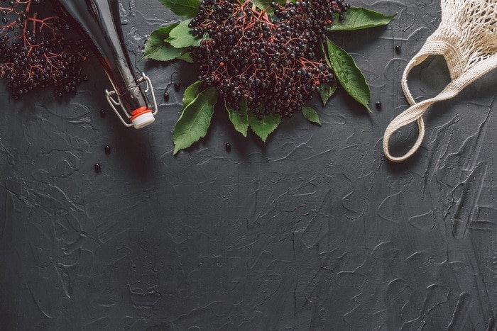 A bottle of elderberry syrup on a dark background with fresh elderberries and string bag.